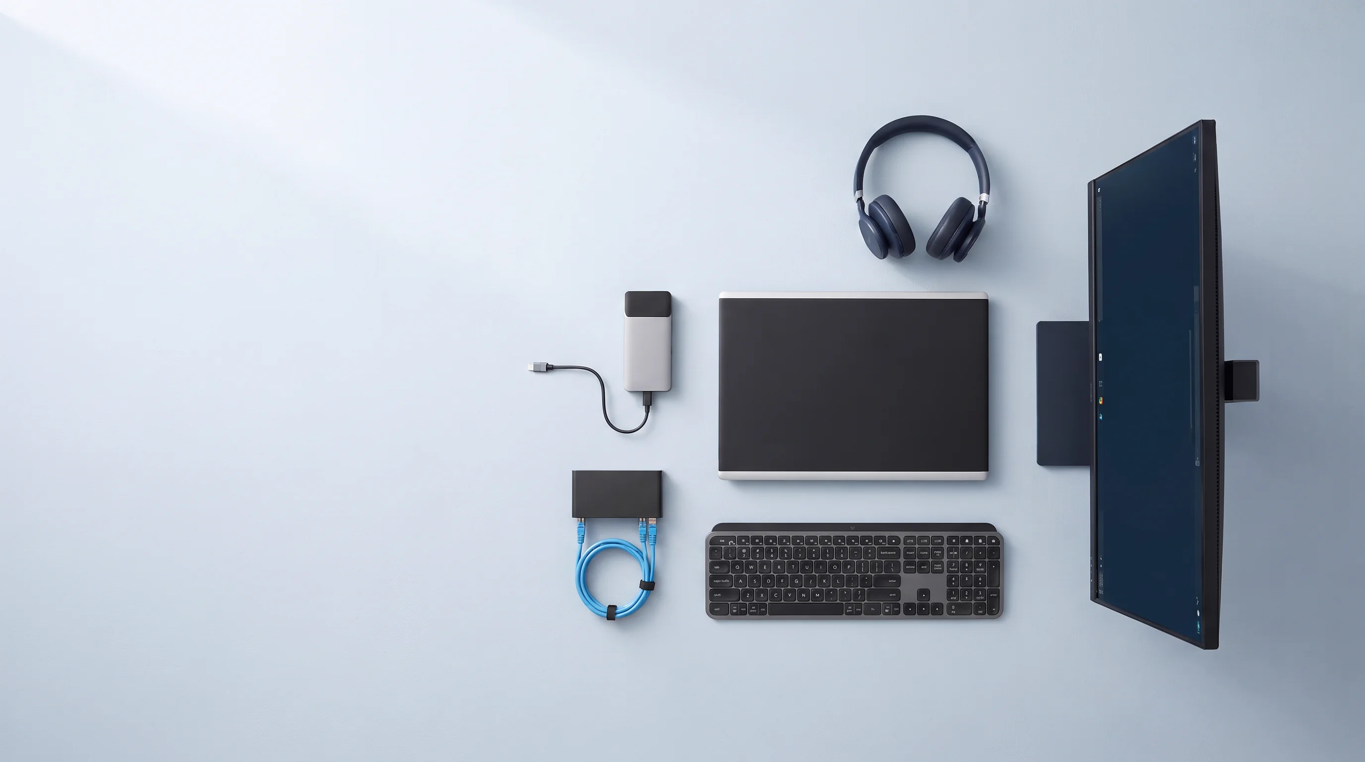 Laptop, monitor, keyboard, headset, dock and network switch laid out on a pale blue surface